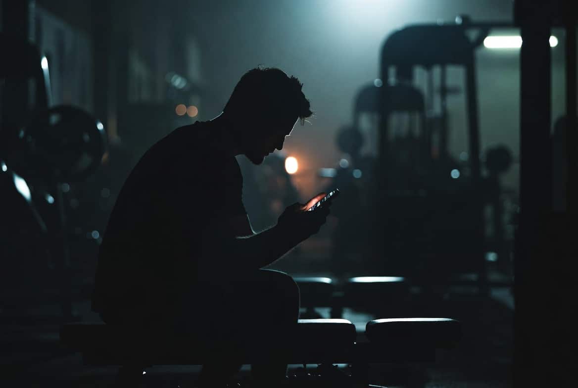 Fitness coach sitting in shadow at night, face lit only by phone screen, scrolling through engagement numbers — visual metaphor for posting without conversion
